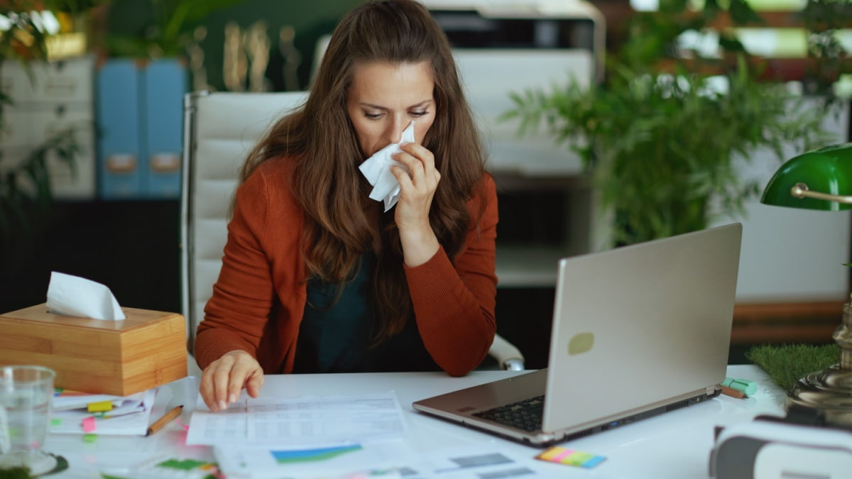 Een vrouw met een zakdoek aan haar neus werkt aan haar bureau achter een laptop.