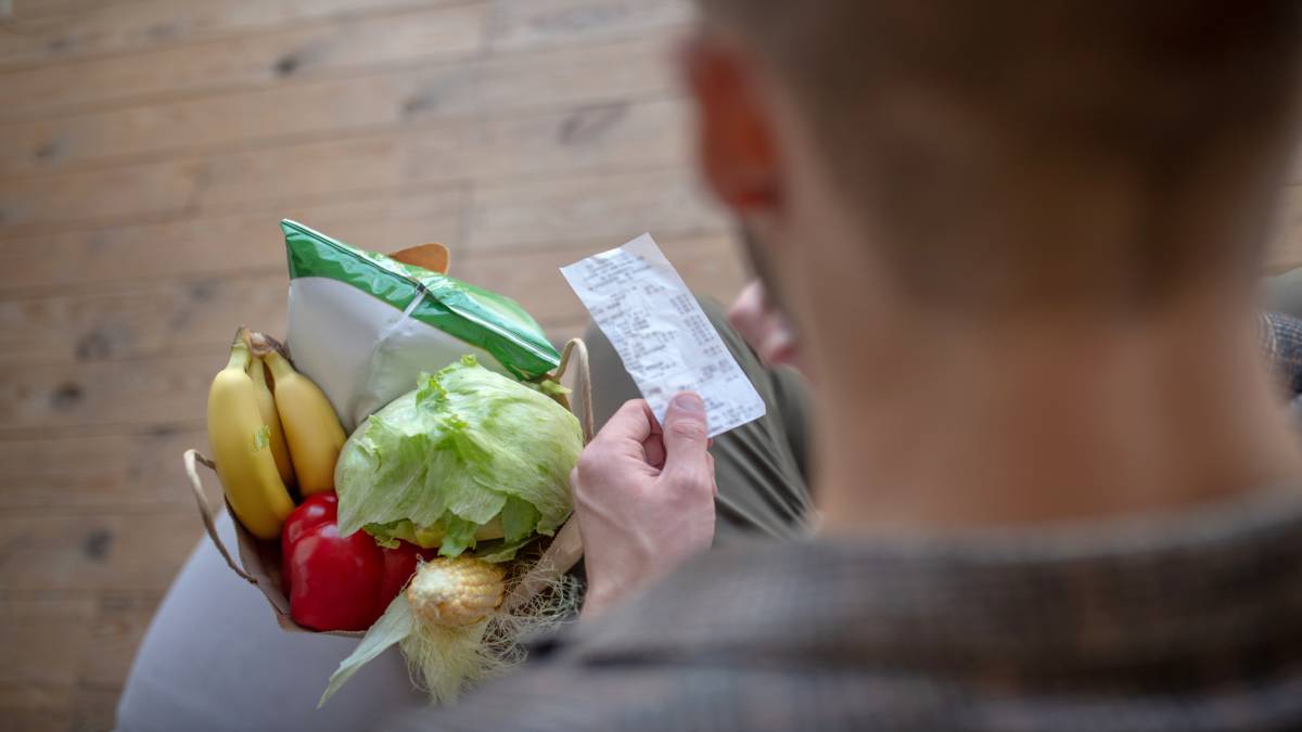 Een man kijkt naar een kassaticket, met een tas vol groenten en bananen op schoot.