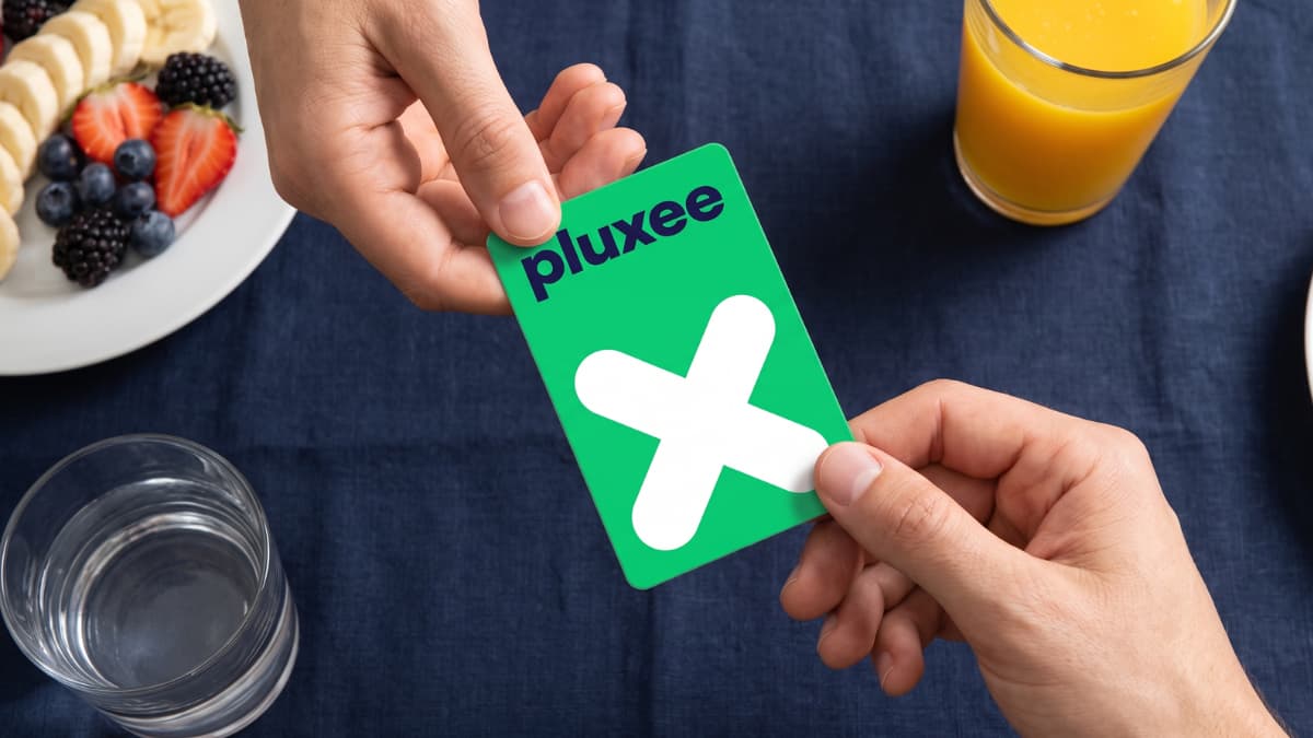 Een hand overhandigt een groene Pluxee kaart met wit kruislogo aan een andere persoon boven een donkerblauw tafelkleed met een bord fruit en glazen sinaasappelsap en water.