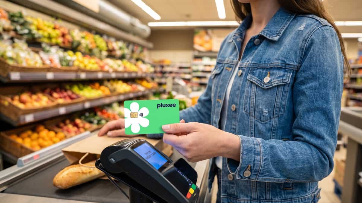 Een vrouw in een jeansjasje houdt een groene Pluxeekaart met witte bloem en gouden chip boven een betaalterminal aan de kassa van een supermarkt met verse groenten op de achtergrond.